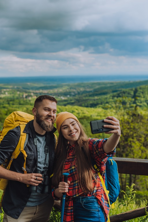 Couple of young hikers taking selfie with a smartphone while wearing camping backpacks and binocular and standing on a lookout