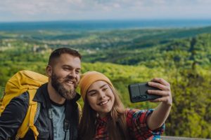 Couple of young hikers taking selfie with a smartphone while wearing camping backpacks and binocular and standing on a lookout